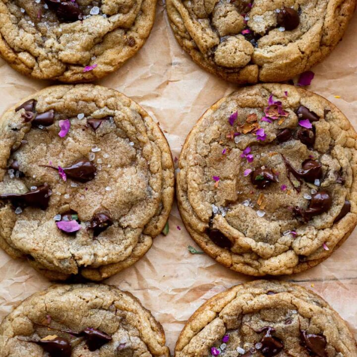 overhead image of 6 round chocolate chip cookies sprinkled with pink rose petals and sea salt on brown parchment paper