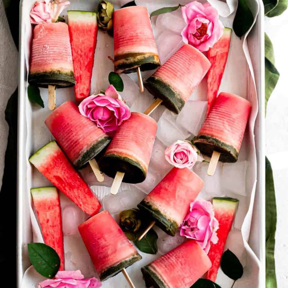 cover image of watermelon mint popsicles an overhead shot of pink popsicles with green on the bottom on a baking tray with pink flowers and ice in the tray