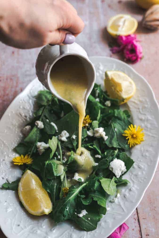 45 degree angle shot of dandelion dressing being poured onto simple greens salad with a wedge of lemon and sprinkled with goat cheese