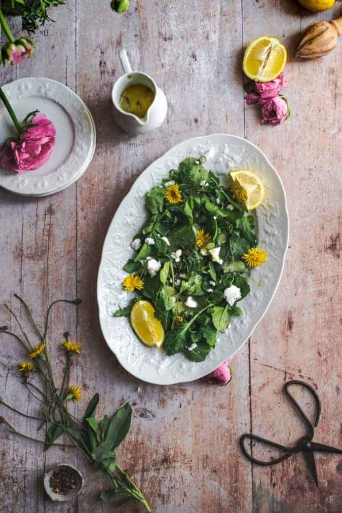 overhead image of a vintage white serving bowl filled with dandelion greens and arugula, lemon wedges, and sprinkled with goat cheese on a wood rustic surface surrounded with wild dandelions and other flowers