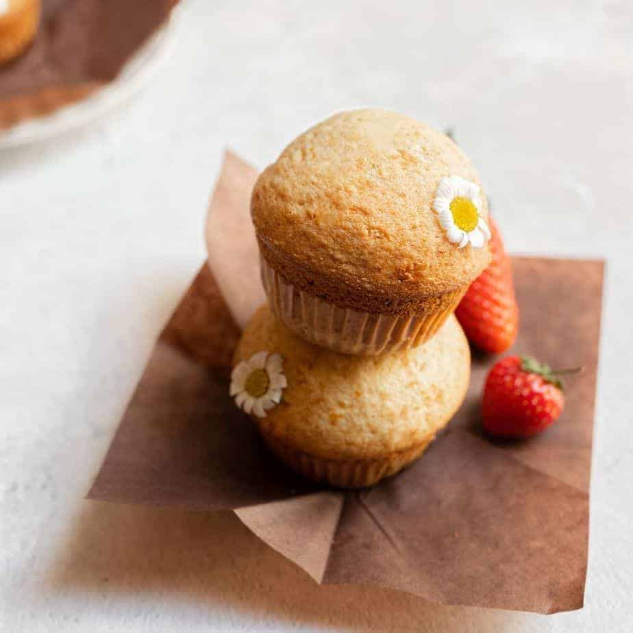 cover image two strawberry muffins on brown muffin liner garnished with a chamomile flower on a bright surface