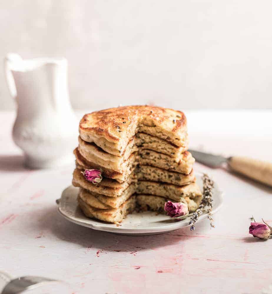 close up shot of a stack of pancakes wiith rose buds and lavender petals on a pink 