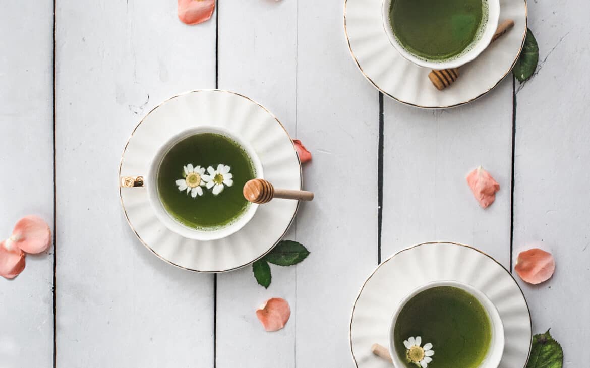 overhead shot of tea cups and saucers with bright green matcha tea with dainty chamomile flowers floating in them and pink rose petals