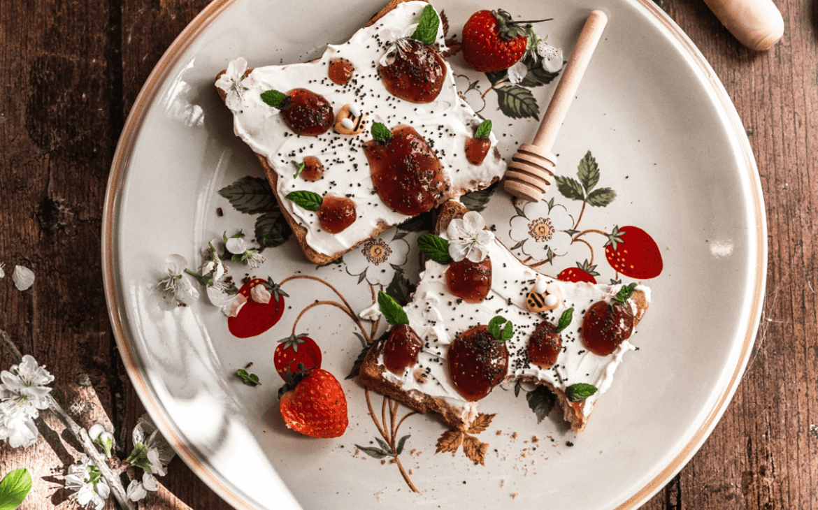 overhead shot of toast covered with mascarpone cheese with dollops of jam shaped like strawberries and mint leaves on a vintage plate with wild strawberries