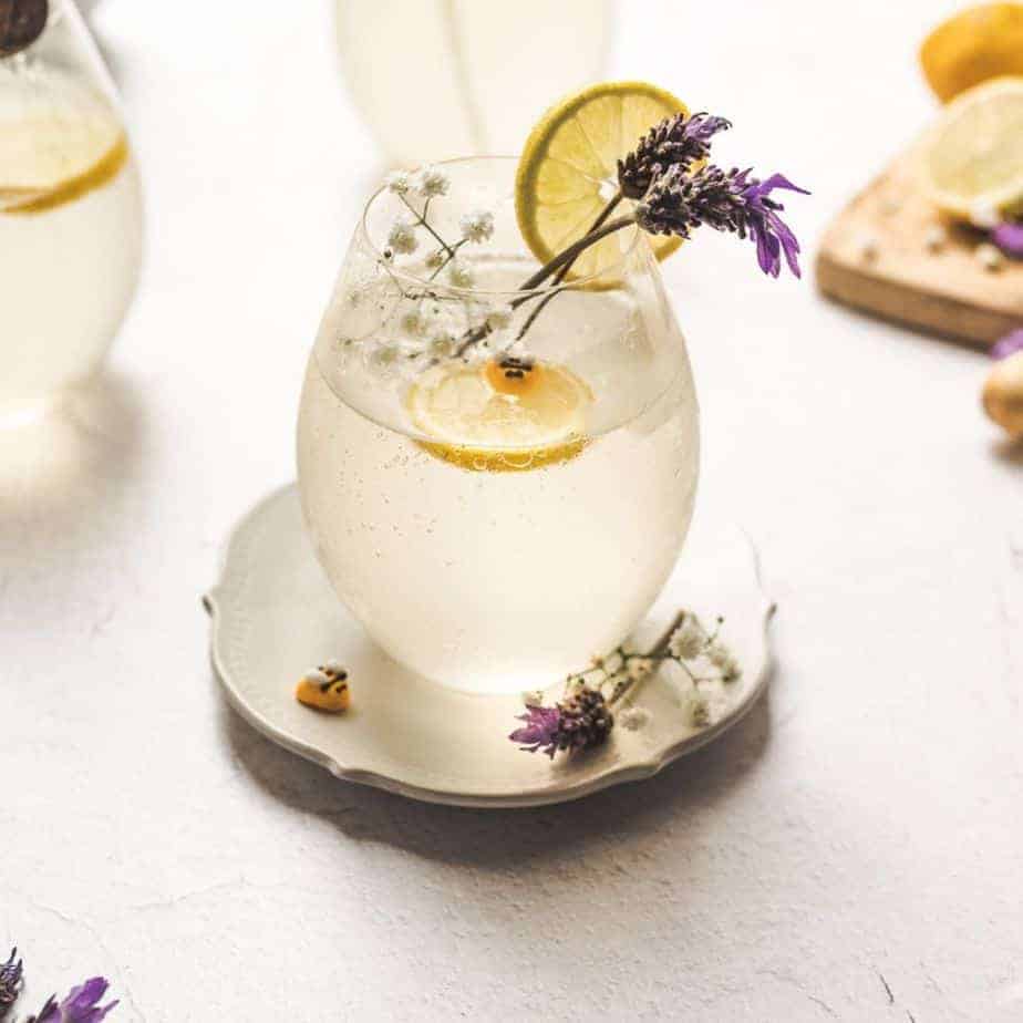 close up shot of cocktail glass filled with lavender honey lemon drink, grarnished with fresh lavender, babies breath flowers, a lemon slice and mini bee sprinkle