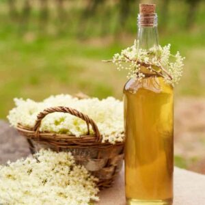 close up image of bottle filled with elderflower syrup with freshly picked elderflowers in a basket to the left of it in front of a grassy field