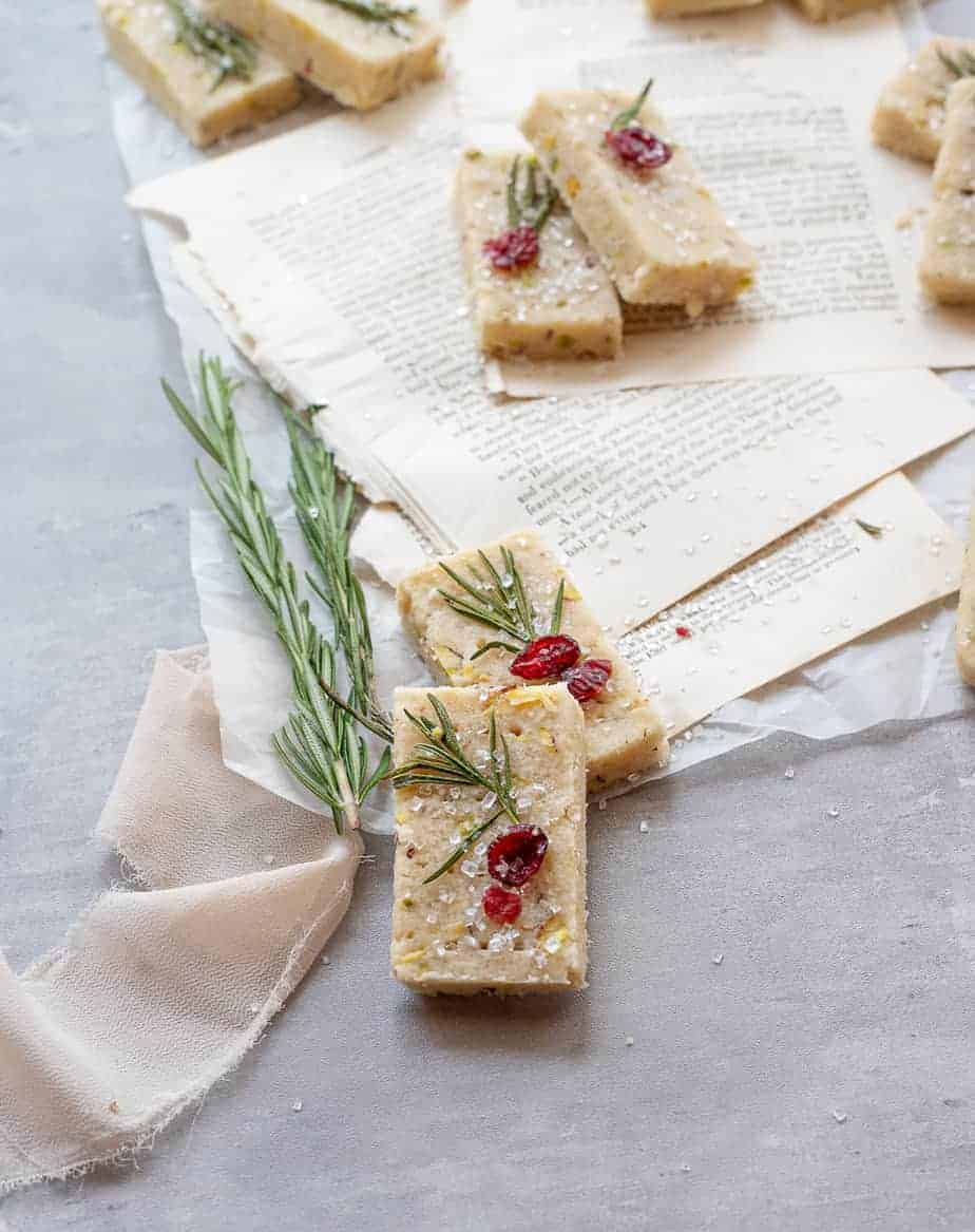 close up 45 degree angle shot of pistachio shortbread - rectangle cookie on concrete background and parchment paper, with a rosemary sprig and cranberry and blush colored ribbon