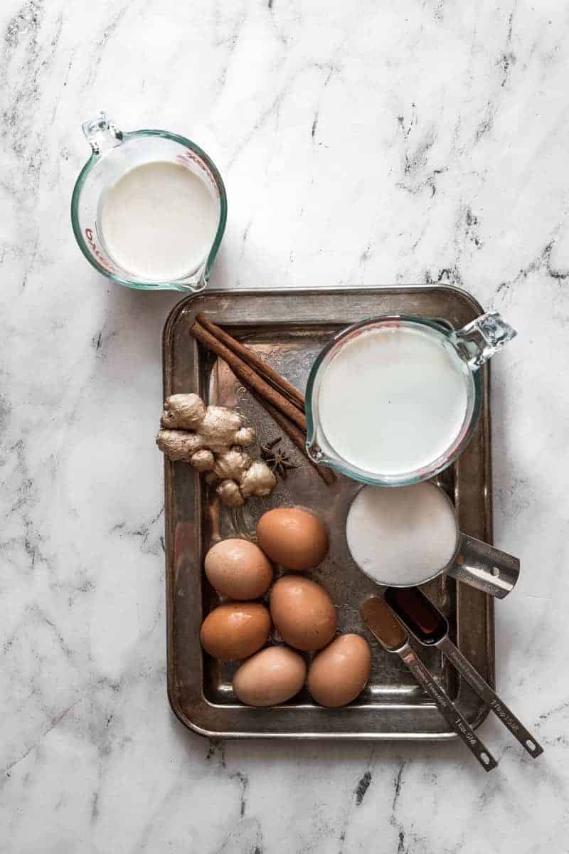 overhead shot of measuring cups with cream, milk, eggs, cinnamon, ginger for eggnog on a white marble background