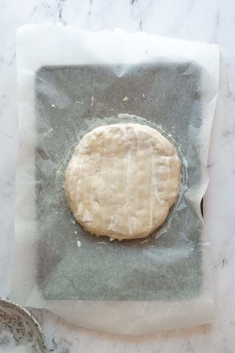 scone dough shaped in a circle on a piece of white parchment paper, brushed with buttermilk