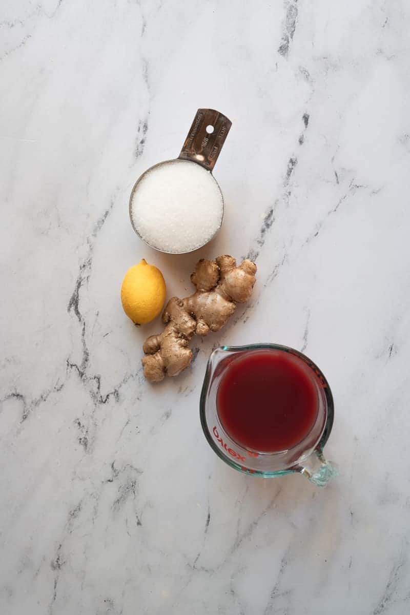 overhead shot of ingredients for pomegranate ginger whisky sour syrup, a cup of sugar, a lemon, a ginger root, and a cup of pomegranate juice on a white marble floor