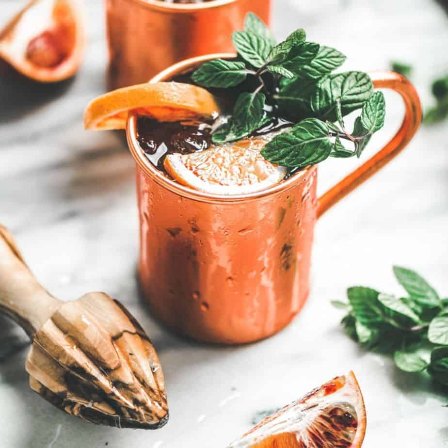 close up image of cold copper mug with blood orange and mint garnish, filled with blood orange Moscow Mule, next to a wooden citrus juicer
