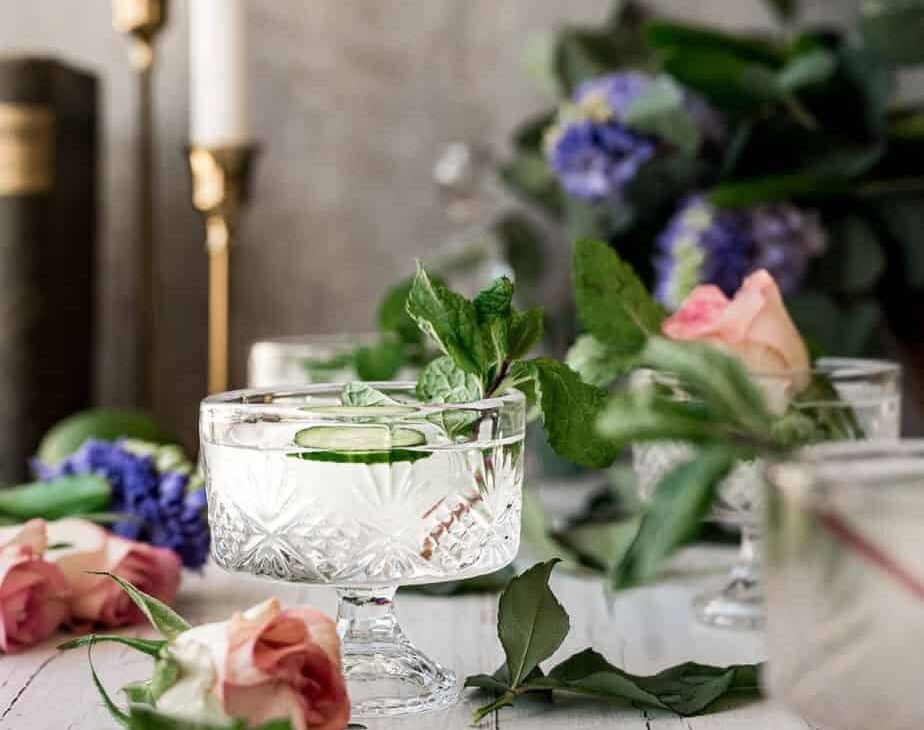 close up image of whisky glass filled with clear cocktail, garnished with cucumber, surrounded by pink roses, purple lilacs, and in front of two golden candle sticks for a cinematic scene