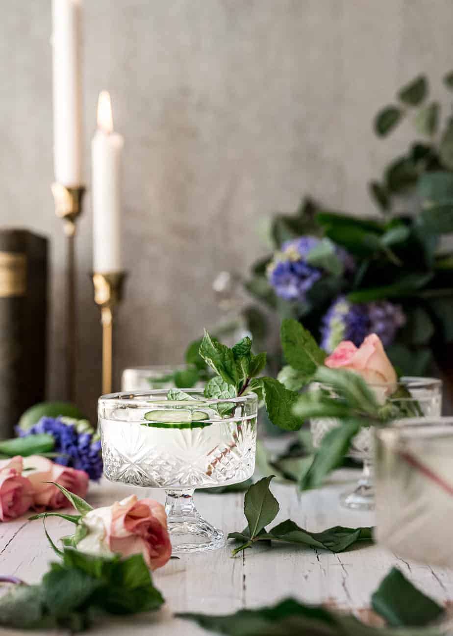 close up shot of small whisky glass with a refreshing cucumber mint garnish, surrounded by pink roses and purple lilacs and candle sticks in the back
