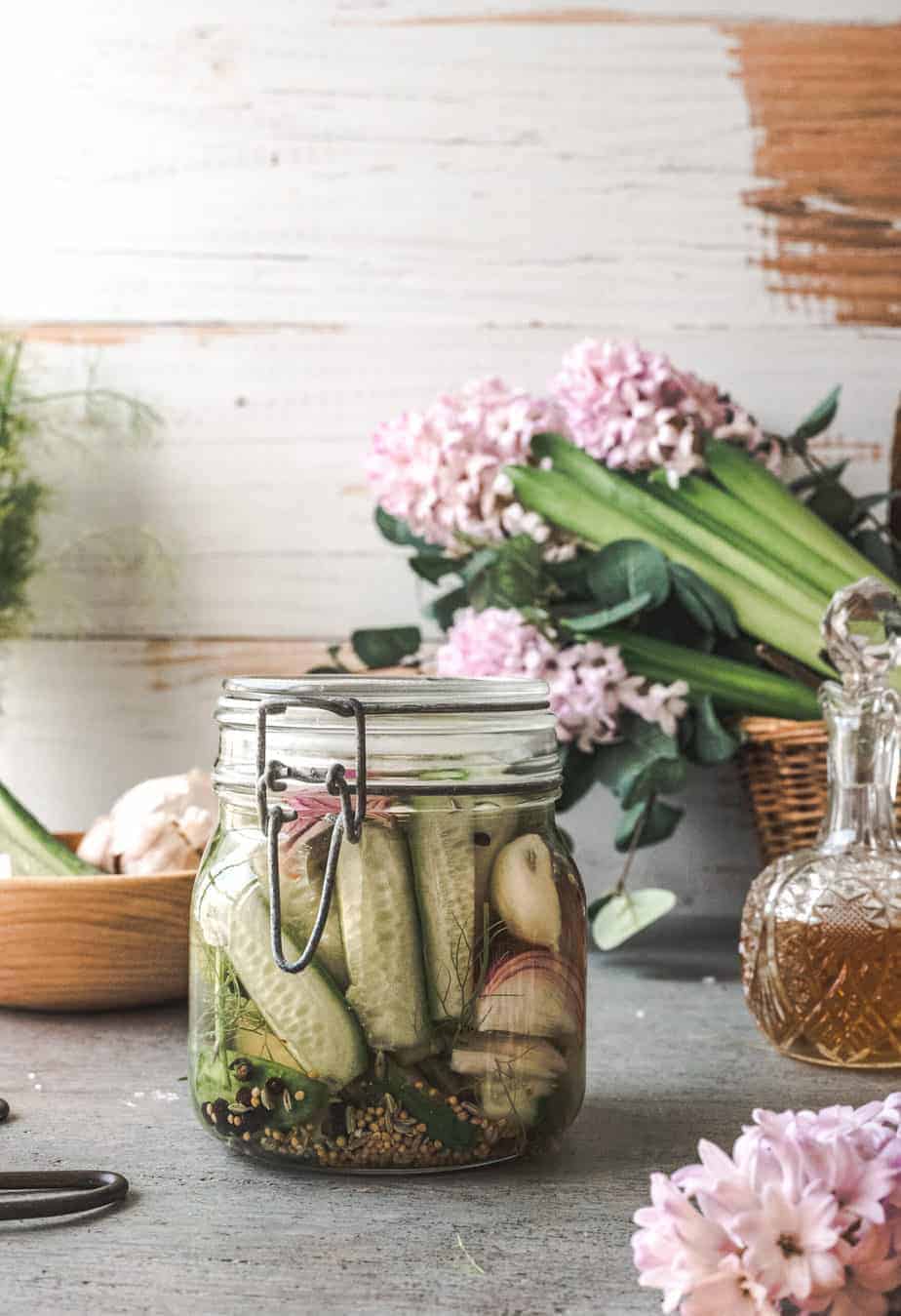 jar of homemade apple cider vinegar pickles - cucumbers with onions, garlic, seeds in a rustic table with apple cider vinegar in a decanter and lavender flowers in a basket in the background