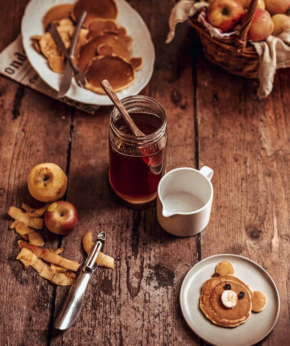 rustic image with a brown wooden table with a basket full of apples, and a white dish with bear shaped pancaked and a jar filled with apple peel pancake syrup with a spoon in it