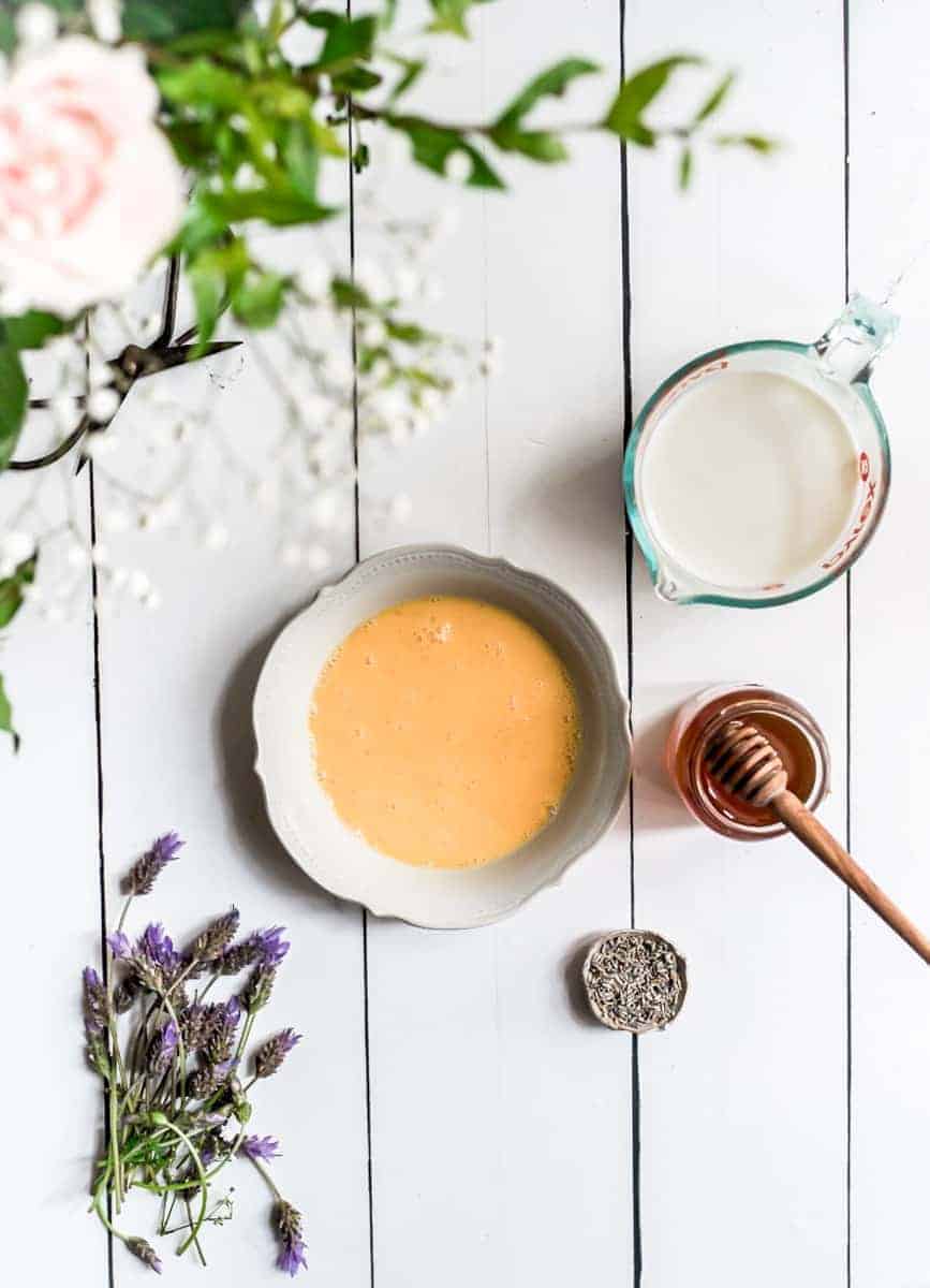 Overhead image of a white wooden table top with the ingredients needed to make lavender honey icecream. egg yolks, honey, lavender, milk and cream