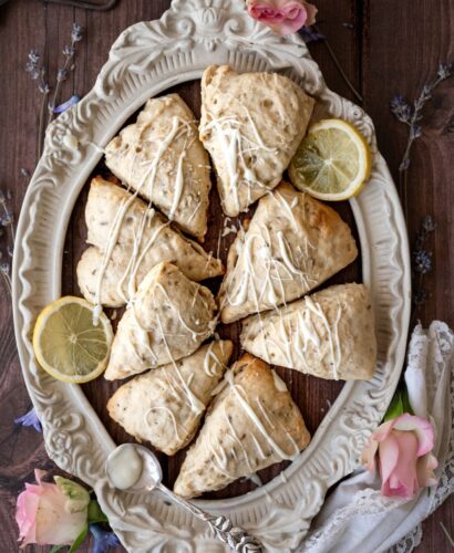 overhead shot of a brown rustic background with a white vintage frame surrounded by dainty flowers: lavender, rose, and lilac, and filled with bright lavender lemon scones, drizzled with white icing