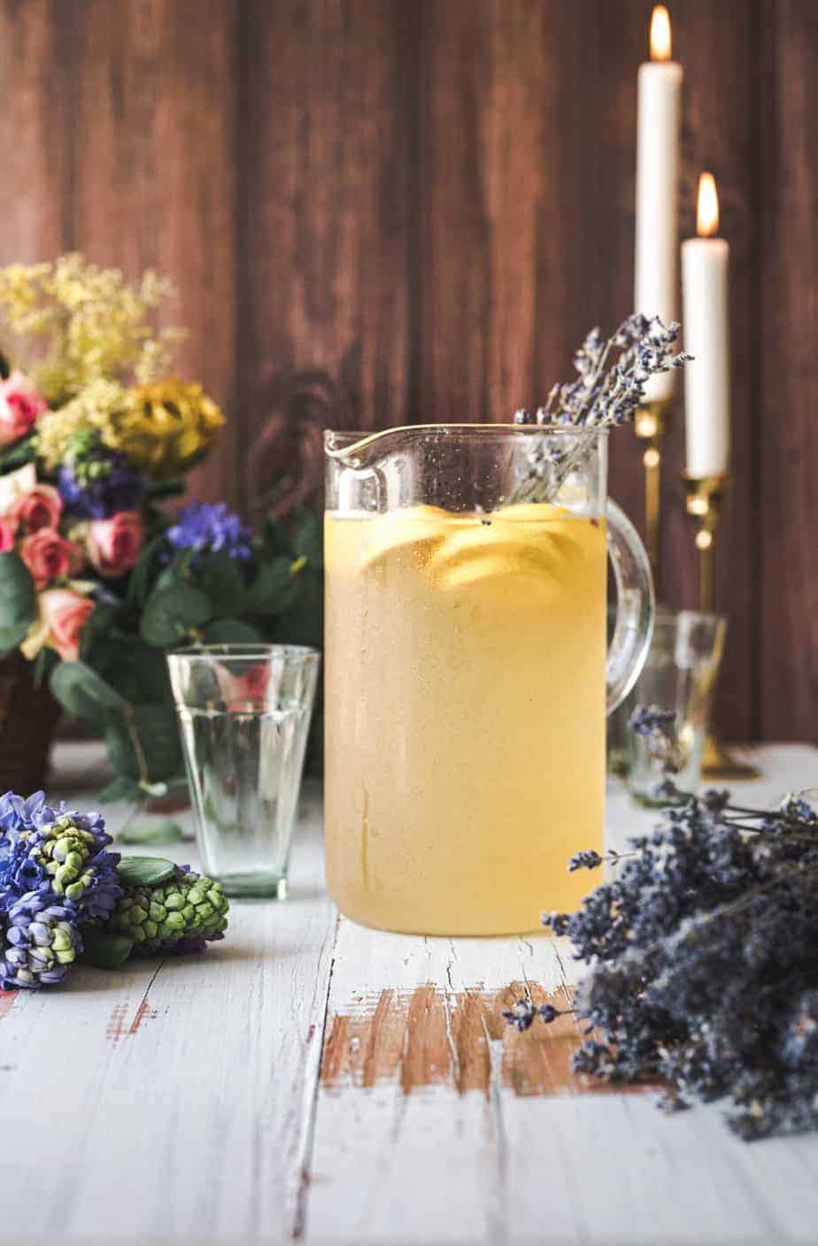 a pitcher filled with lavender lemonade, on a white rustic wooden table top, with a brown wooden panels in the background, with candles lit and a basket of fresh market flowers off to the side