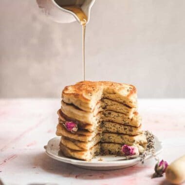close up shot of a stack of pancakes wiith rose buds and lavender petals on a pink surface with woman's hands pouring maple syrup onto the stack