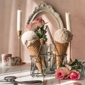 image of vintage glass carrier filled with sugar cones, and pink ice cream scoops, garnished with pink roses, and in front of vintage white frame and two candlle sticks