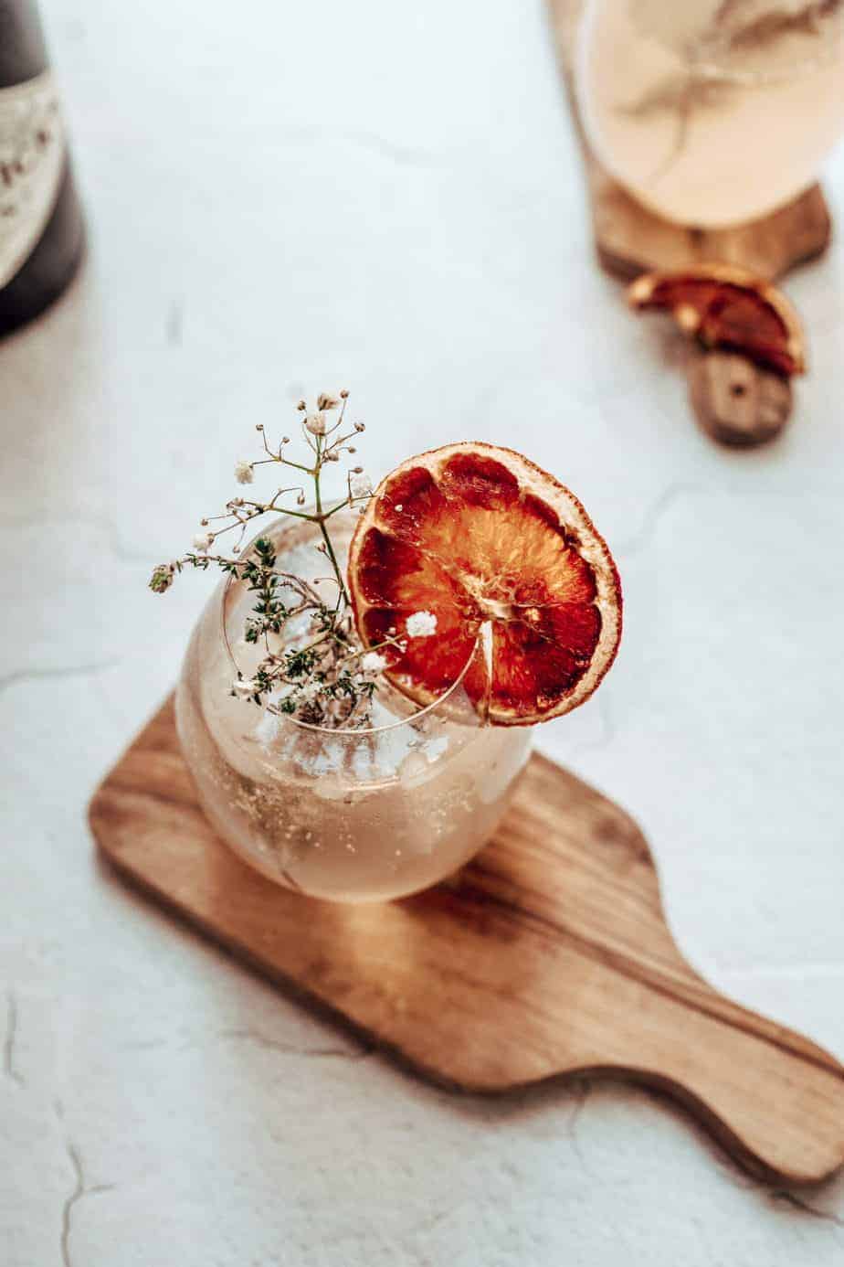 close up shot of a gin and tonic in high ball glass on top of a wooden cutting board, garnished with orange slice and rosemary