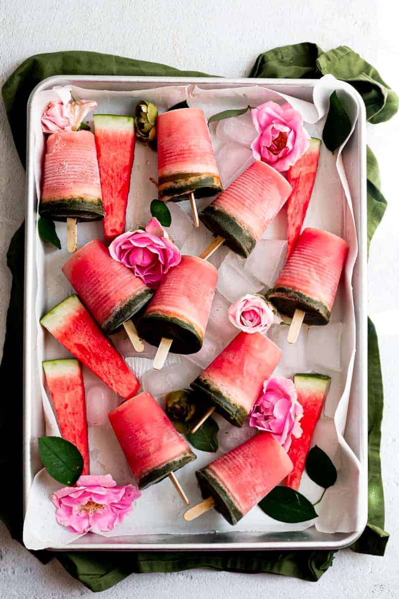 watermelon mint popsicles an overhead shot of pink popsicles with green on the bottom on a baking tray with pink flowers and ice in the tray