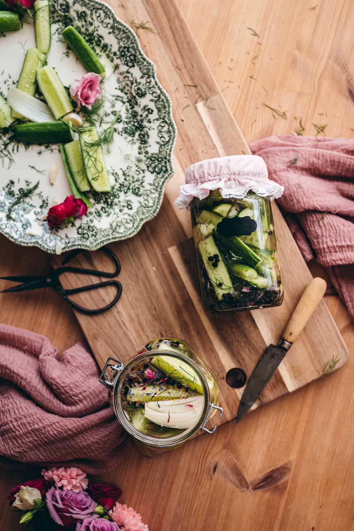 apple cider vinegar pickles cover image, open bottle of pickles on left hand corner of image with dried edible flower petals, sliced cucumbers and salt. a closed jar of pickles labeled pickles with a pink jar lid cover, and a green vintange victorian style plate with sliced pickles, dill, and pink flowers, next to a pair of scissors and a knife on top of a pink cheesecloth