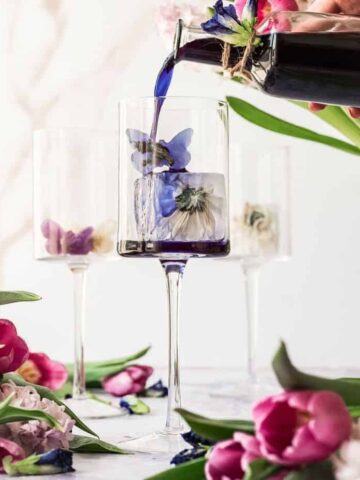 cover image of butterfly pea syrup three glasses with flower icecubes with womans hand with pink nails pouring bright blue butterfly pea syrup into glass 1