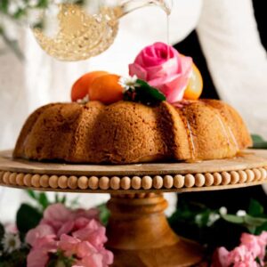 woman pouring orange blossom syrup out of a vintage crystal decanter over a moist olive oil citrus cake, garnished with clementines and pink flowers