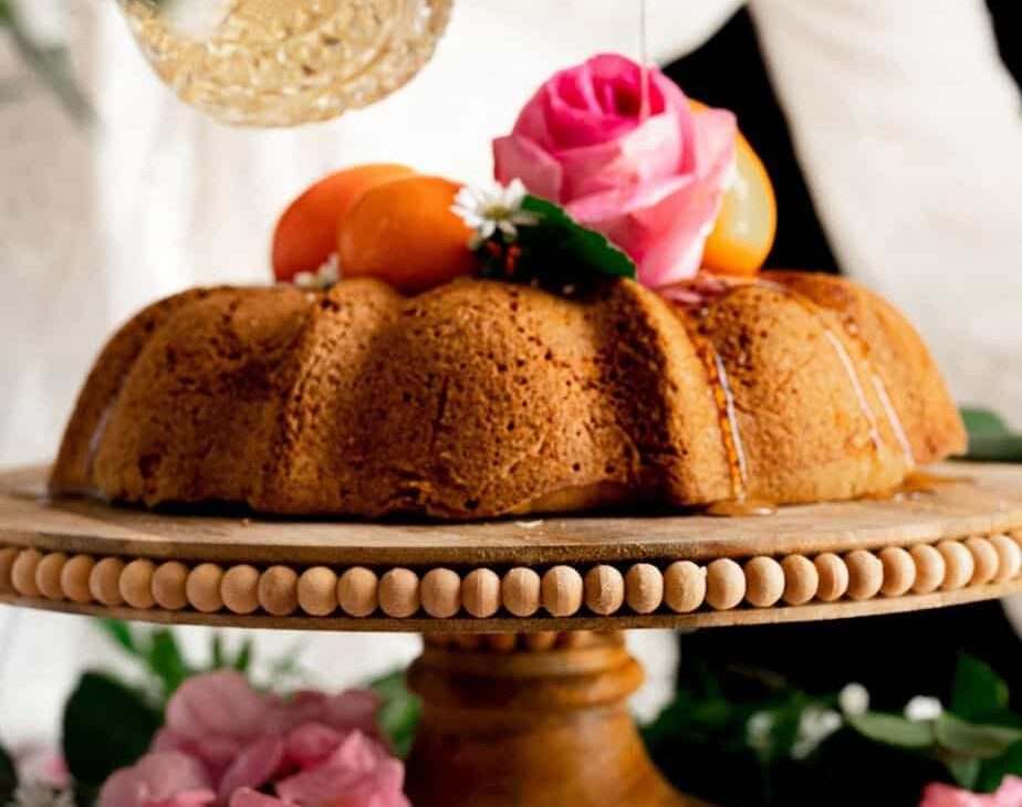 woman pouring orange blossom syrup out of a vintage crystal decanter over a moist olive oil citrus cake, garnished with clementines and pink flowers