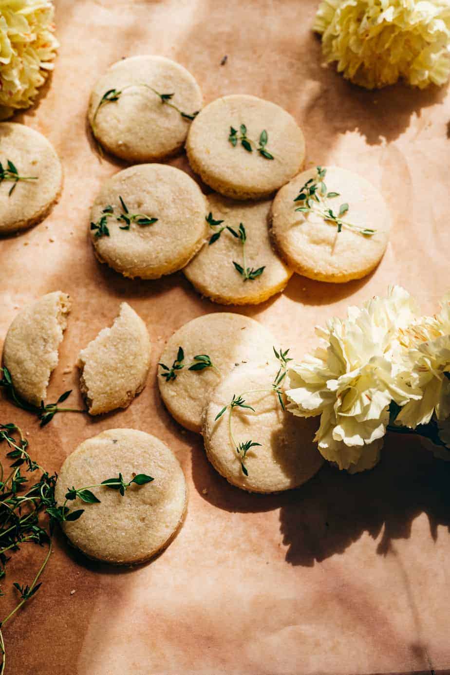 angle view of 10 round cookies, one broken in half all garnished with bright green thyme- with bright sunshine casting dark shadows and yellow flowers styled around the cookies