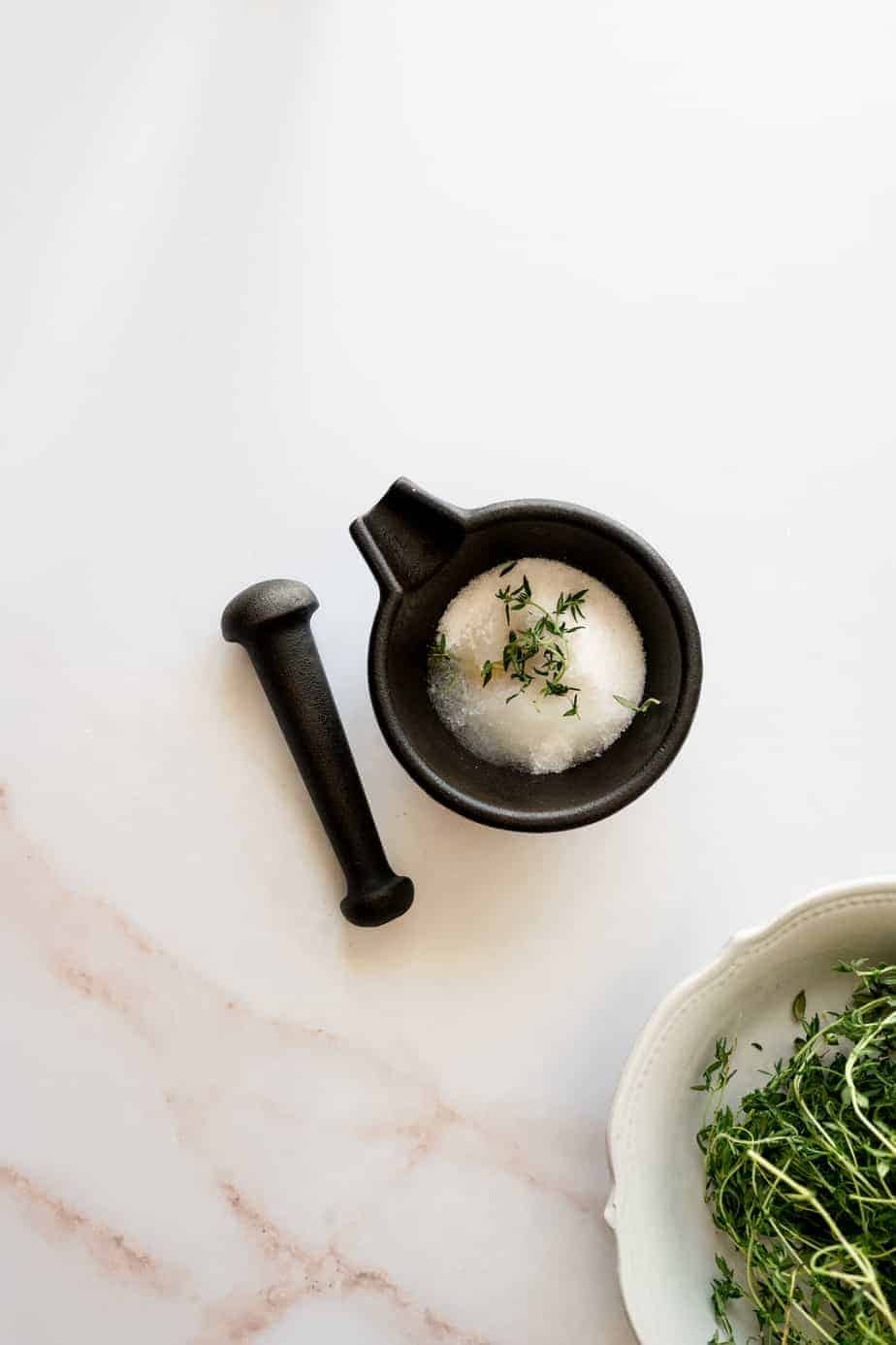 overhead shot of a white marble surface with mortar and pestle, mortar filled with sugar and thyme, and bowl of fresh thyme