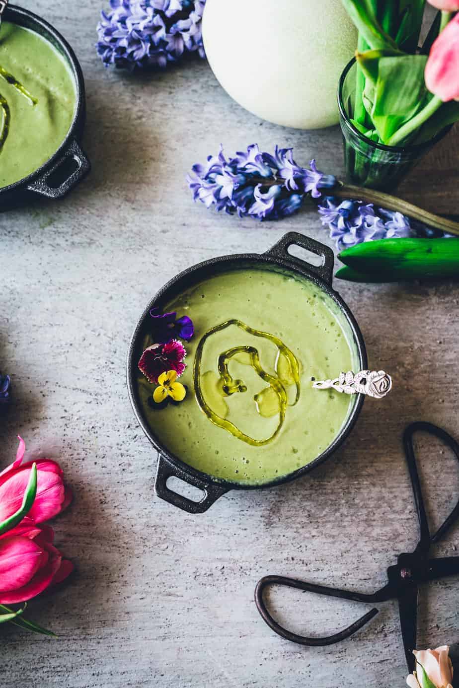overhead shot of vibrant green gazpacho soup drizzled with olive oil and garnished with edible flowers with spoon in bowl