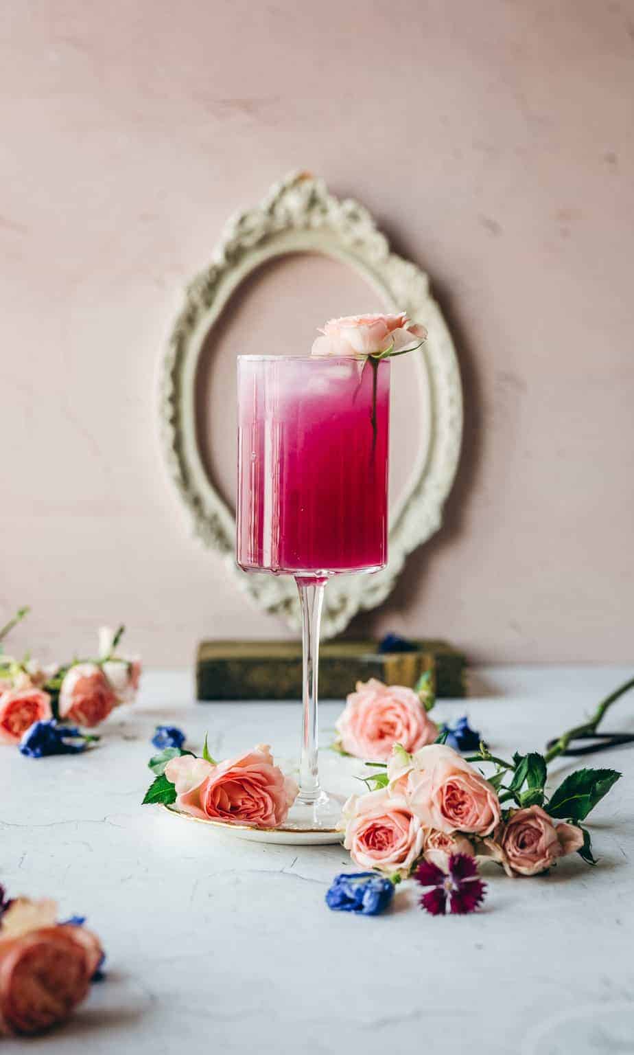 image of tall cocktail glass with vibrant pink cocktail surrounded by pink roses and butterfly pea flowers