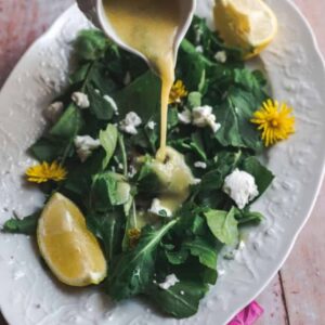 45 degree angle shot of dandelion dressing being poured onto simple greens salad with a wedge of lemon and sprinkled with goat cheese