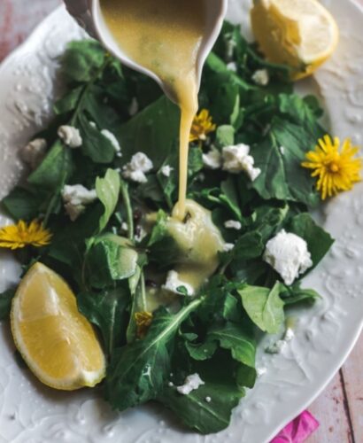 45 degree angle shot of dandelion dressing being poured onto simple greens salad with a wedge of lemon and sprinkled with goat cheese