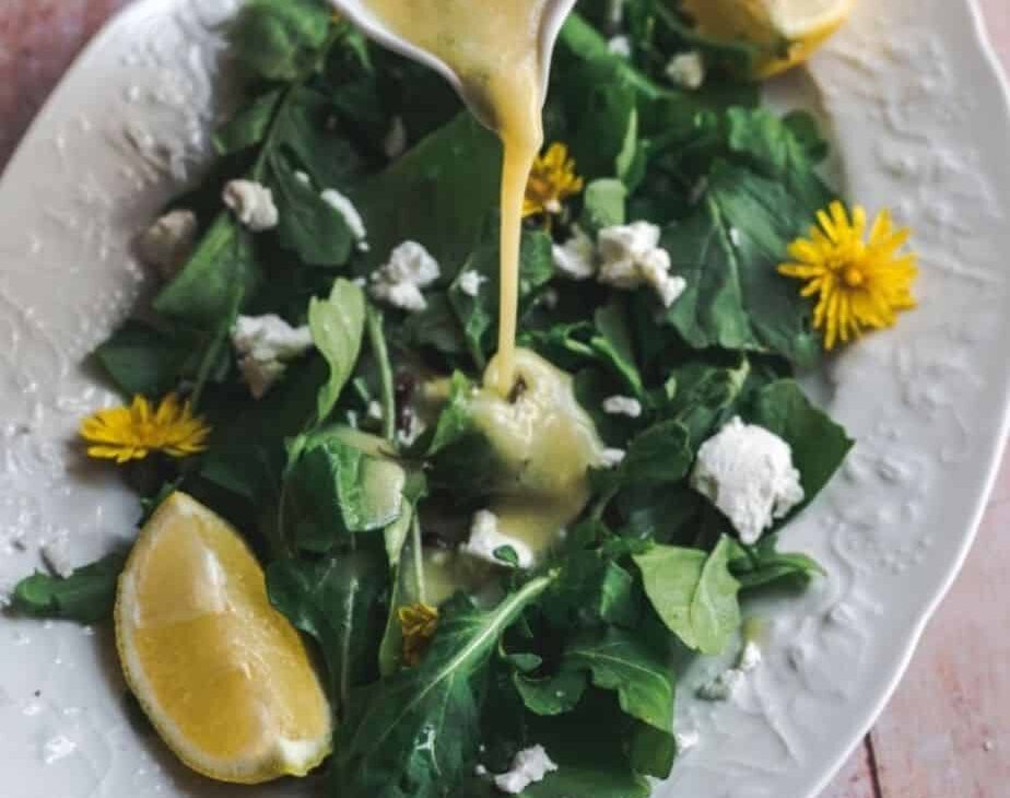 45 degree angle shot of dandelion dressing being poured onto simple greens salad with a wedge of lemon and sprinkled with goat cheese