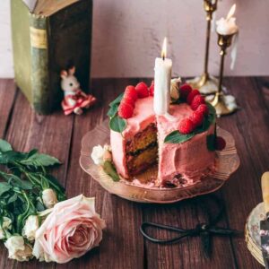 raspberry chamomile cake pink cake on pink vintage plate with a large candle on top, garnished with fresh raspberry slices and basil leaves on a brown rustic table