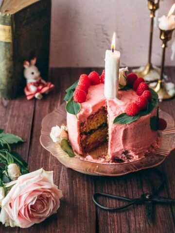 raspberry chamomile cake pink cake on pink vintage plate with a large candle on top, garnished with fresh raspberry slices and basil leaves on a brown rustic table