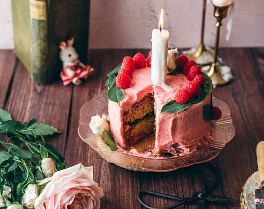raspberry chamomile cake pink cake on pink vintage plate with a large candle on top, garnished with fresh raspberry slices and basil leaves on a brown rustic table