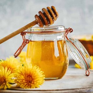 dandelion honey close up in a jar with olive wood honey spoon surrounded by dandelions