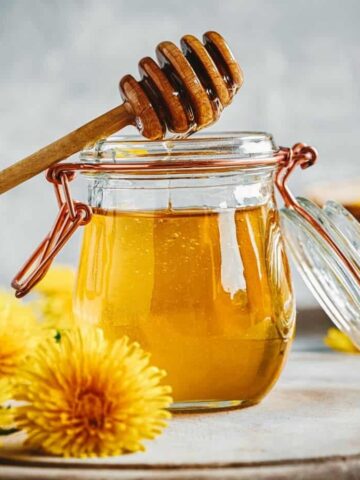 dandelion honey close up in a jar with olive wood honey spoon surrounded by dandelions