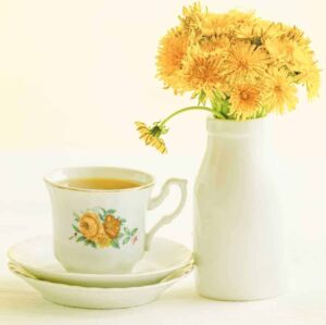 image of vintage tea cup with yellow flowers with yellow tea on a saucer and a small white vase filled with dandelions