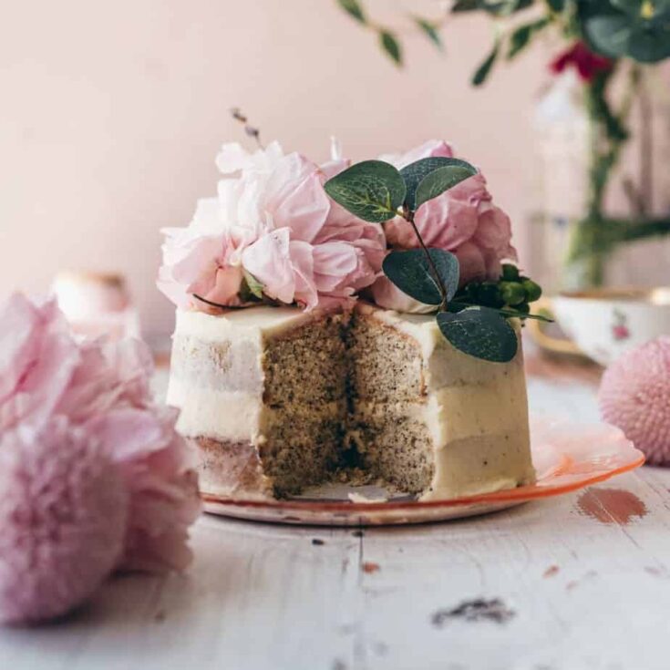 close up image of a small frosted cake sliced in the middle to show tea infused layers topped with pink peonies with a pink background