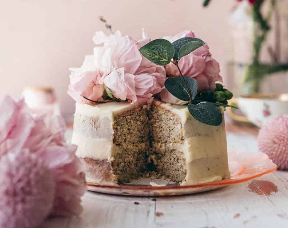 close up image of a small frosted cake sliced in the middle to show tea infused layers topped with pink peonies with a pink background