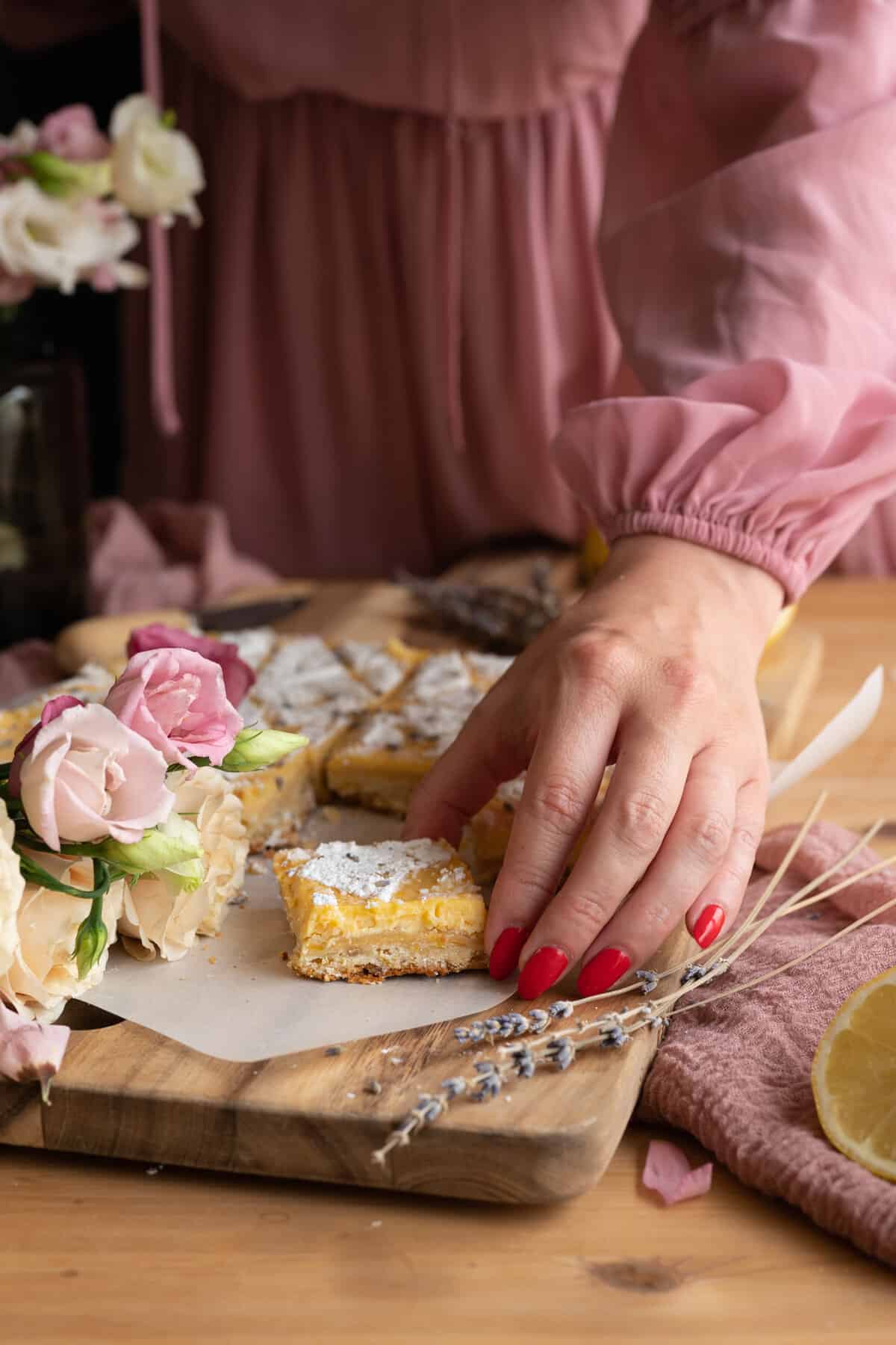 womans hand with red nails grabbing lavender lemon bar, next to a pink bouqet of roses