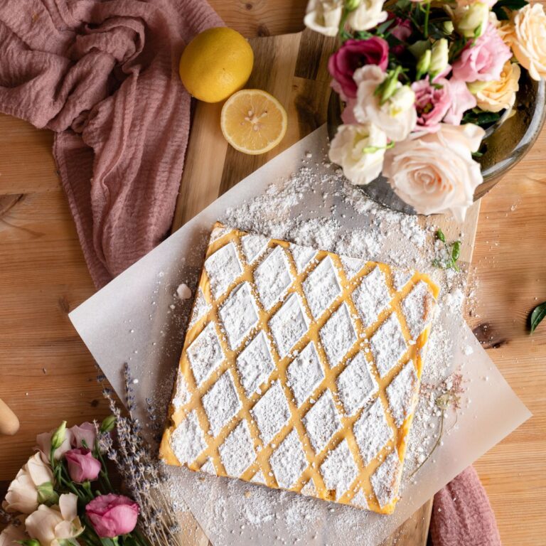 overhead image of lavender lemon bars with diagnol pattern of powdered sugar on rectangle slice of parchment paper on a rustic table next to roses