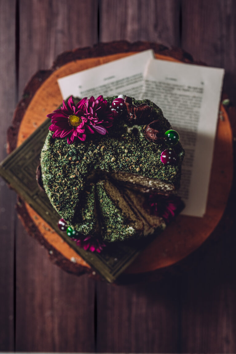 overhead shot of a small round cake covered in edible green moss, on top of a wood log cake stand with rustic pages from a book and magenta flowers
