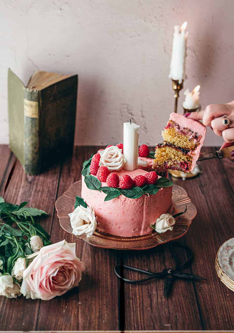 slice of pink frosted cake topped with raspberries and basil leaves with woman's hand pulling a slice of cake to serve