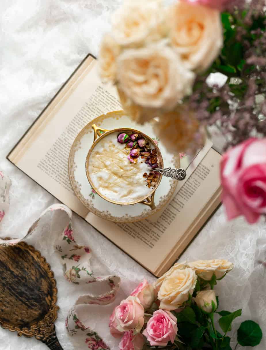 overhead image of vintage tea cup on vintage book filled with frothed rose milk, drizzled with golden honey and garnished with pink rose bud petals next to a bouquet of fresh roses